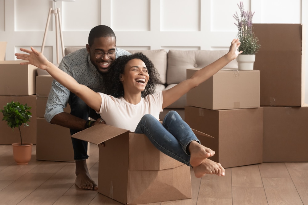 Happy african american young couple first time home buyers having fun unpacking laughing on moving day, excited wife riding sitting in cardboard box while black husband push it in new house apartment